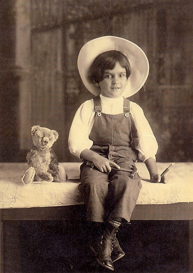 Historic black and white photo of a young Frida Kahlo at age 6 in 1913, captured by photographer Guillermo Kahlo, sitting confidently with a teddy bear and toy in hand, showcasing early signs of the formidable artist she would become.