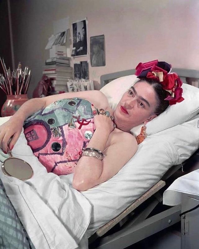 Colorized photograph of artist Frida Kahlo reclining on a bed with a painted plaster corset, adorned with vibrant flowers in her hair, exemplifying her artistic spirit even in times of convalescence.