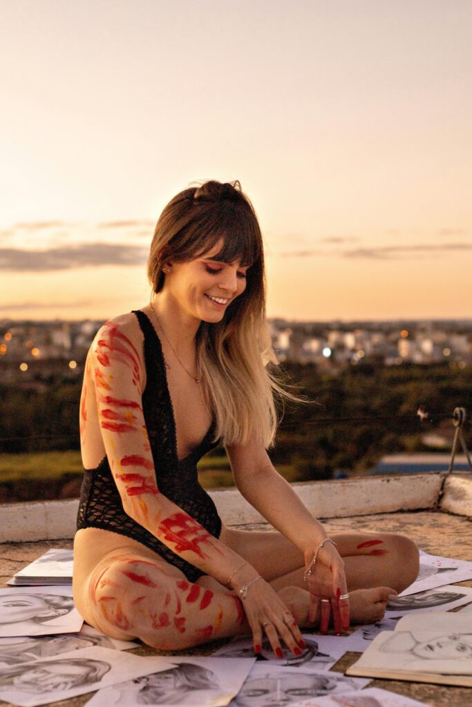Smiling artist with paint-adorned arms sitting among sketches at sunset, embracing a new environment for creative inspiration.