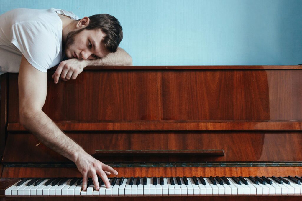 Despondent musician resting his head on a piano, fingers lightly touching the keys, depicting the shared struggle of art block among artists.