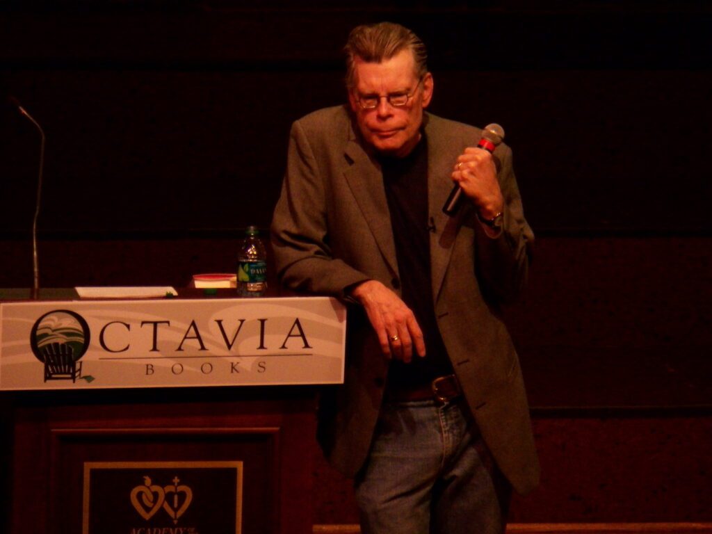 Stephen King speaking at an event at Octavia Books, holding a microphone and leaning on a podium.
