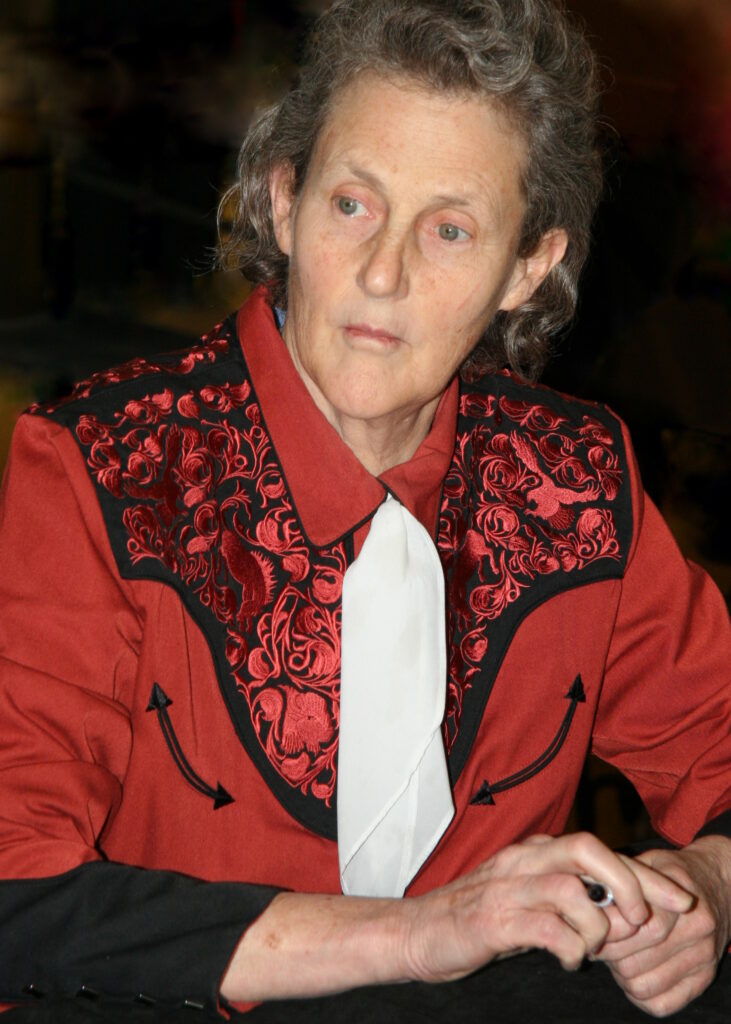 Temple Grandin, renowned author and autism advocate, wearing a red and black embroidered shirt with a white tie, looking thoughtful.