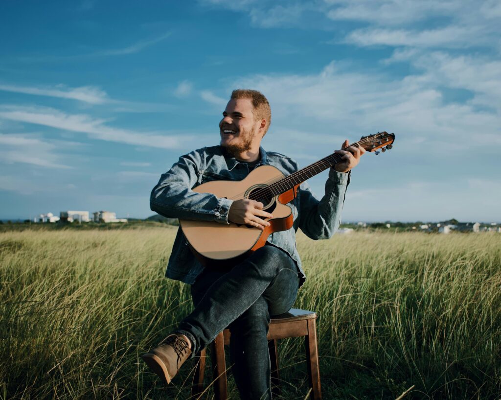 Man joyfully playing acoustic guitar in a grassy field with blue sky background
